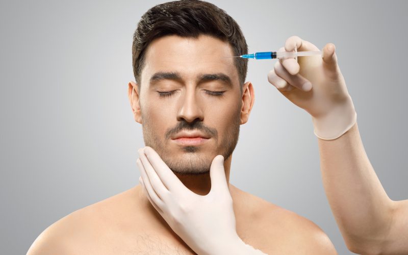 Young man isolated on gray background receiving botox injection from beauty doctor in sterile gloves in forehead to reduce aging, eyes closed